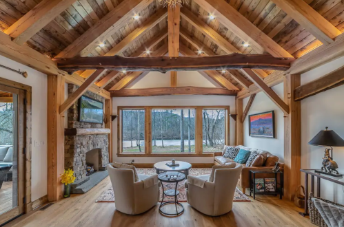 Cozy living room with wooden beams, stone fireplace, large windows, and comfortable seating overlooking a scenic view.