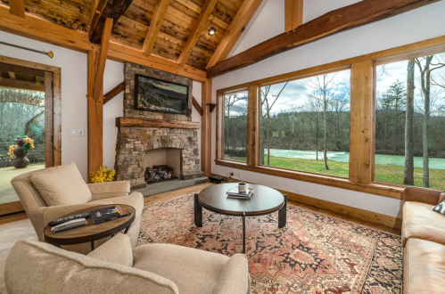 Cozy living room with a stone fireplace, large windows, and rustic wooden beams, overlooking a serene outdoor view.