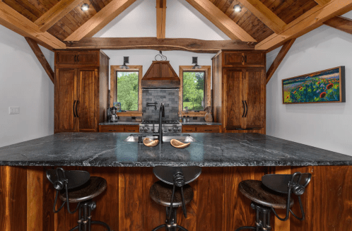 A rustic kitchen with wooden beams, dark countertops, and bar stools, featuring a stove and large windows.