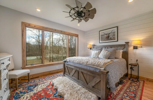 Cozy bedroom with a large window, wooden bed, decorative rug, and soft lighting, featuring a rustic design.