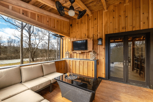 Cozy wooden porch with a sectional sofa, coffee table, and TV, overlooking a serene outdoor view.