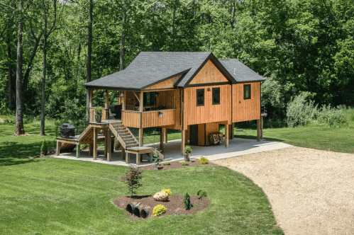 A modern wooden house on stilts surrounded by lush greenery and a gravel driveway.