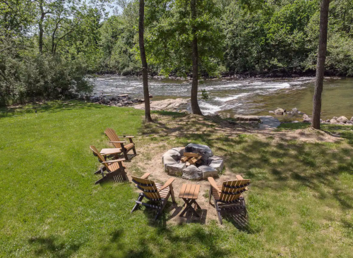A serene riverside scene featuring a fire pit surrounded by wooden chairs and lush greenery.