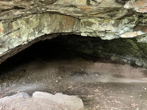 A dark cave entrance with rocky walls and a dirt floor, surrounded by natural stone formations.