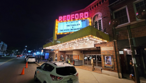 A brightly lit theater marquee reads "Redford" with upcoming shows, surrounded by cars and people at night.