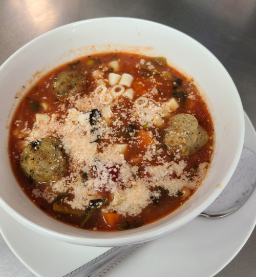 A bowl of soup with meatballs, pasta, vegetables, and grated cheese, served on a white plate with a spoon.