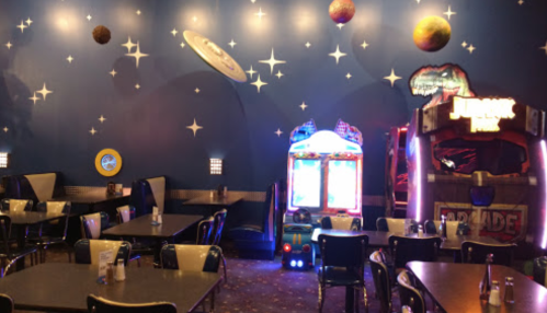 A colorful arcade area with tables, games, and a starry wall mural featuring planets and space themes.