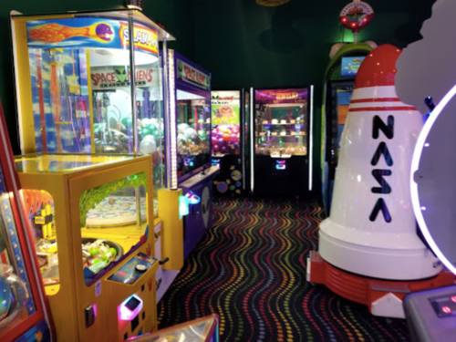 A colorful arcade with various games, including a NASA-themed rocket and claw machines, set against a vibrant carpet.