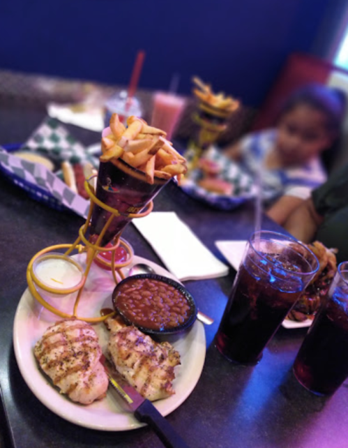 A plate with grilled chicken, baked beans, and a tall serving of fries, with drinks in the background.