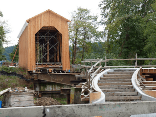 Construction site featuring a wooden structure resembling a covered bridge, with scaffolding and a curved concrete path.