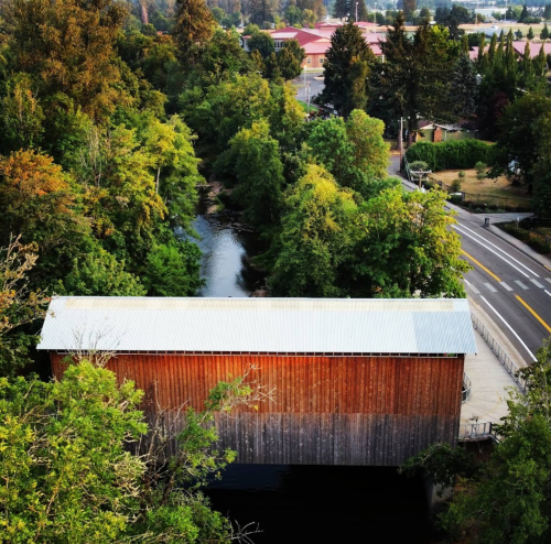 Aerial view of a wooden covered bridge over a creek, surrounded by lush greenery and a road nearby.