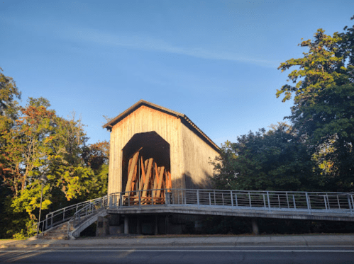 A wooden covered bridge surrounded by trees, with a ramp leading up to its entrance. Clear blue sky above.