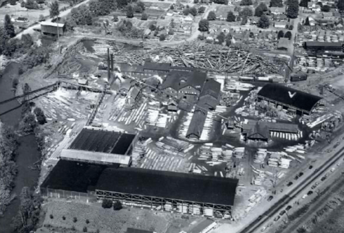Aerial view of an industrial site with buildings and stacked materials near a river, surrounded by greenery.