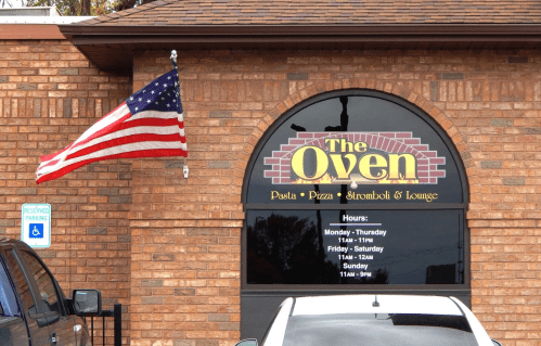 Exterior of The Oven restaurant with an American flag, featuring hours of operation on a brick wall.