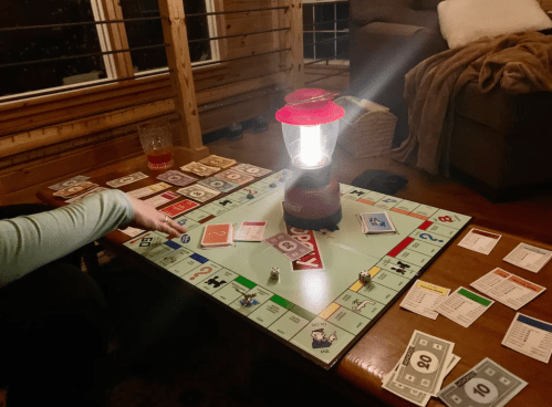 A cozy Monopoly game setup with a lantern, cards, and money on a wooden table in a warmly lit room.