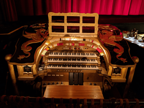 A vintage theater organ with ornate decorations and multiple keyboards, set against a red curtain backdrop.