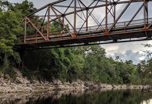 Rusty metal bridge spanning a calm river, surrounded by lush greenery and rocky riverbanks under a cloudy sky.