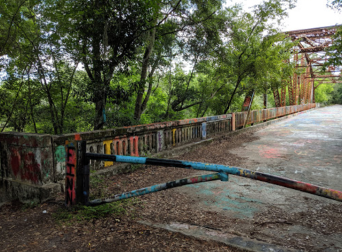 An old, colorful bridge with graffiti, surrounded by lush greenery and a partially closed gate.