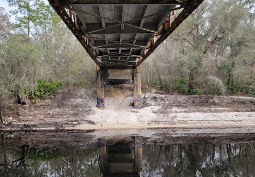 A weathered bridge spans over a calm river, reflecting its structure in the water below, surrounded by lush greenery.