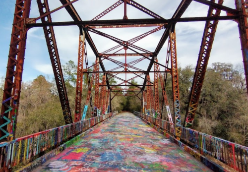 A colorful, graffiti-covered bridge with rusted metal beams, surrounded by trees and a blue sky.