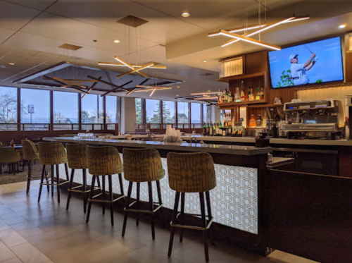 Modern bar area with illuminated counter, high stools, and a TV showing a golfer, surrounded by large windows.
