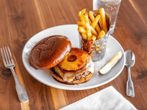 A plate with a burger topped with pineapple, cheese, and fries in a cup, alongside a pickle and a glass of water.