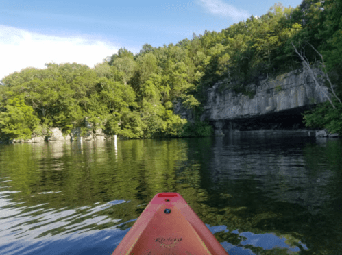 A kayak on calm water, surrounded by lush green trees and rocky cliffs under a clear blue sky.