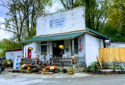 A quaint country store with a porch, decorated with pumpkins and flowers, surrounded by trees in autumn.