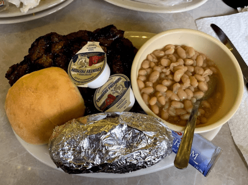 A plate with grilled meat, a roll, baked beans, a foil-wrapped potato, and butter packets.