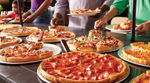 A variety of pizzas on display, with people serving slices at a buffet-style setting.