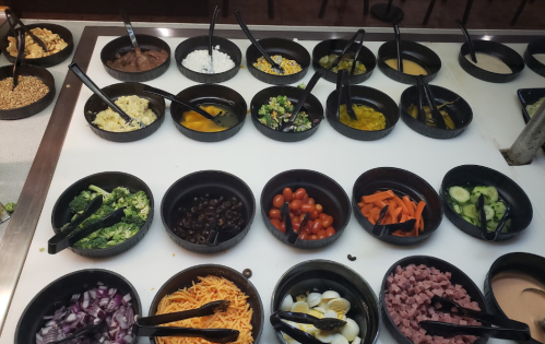 A variety of salad toppings in black bowls, including vegetables, cheeses, and dressings, with serving tongs.