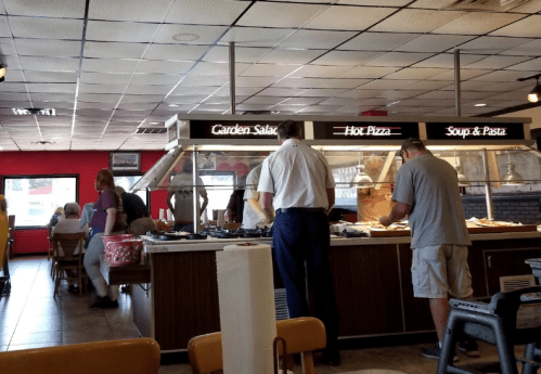 A buffet restaurant scene with diners at a salad and pizza bar, and a few people seated at tables.