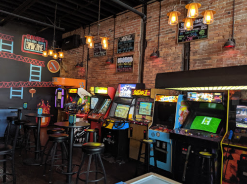 A retro arcade with colorful game machines, bar stools, and exposed brick walls, illuminated by hanging lights.