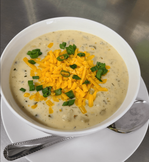 A bowl of creamy soup topped with shredded cheddar cheese and chopped green onions, served with a spoon on the side.