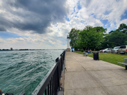 A waterfront view with a path, trees, and a lighthouse in the distance under a cloudy sky.