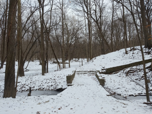 A snowy landscape with bare trees, a path leading to a stone bridge over a small creek in a serene park setting.