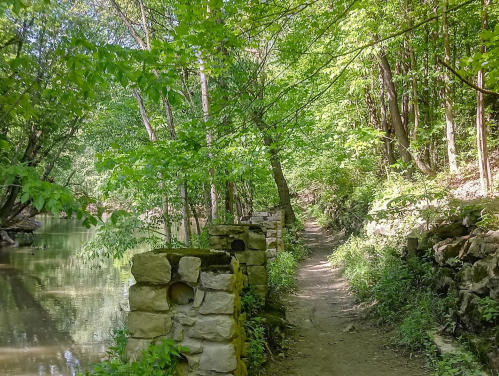 A serene path lined with stone walls, surrounded by lush green trees and a calm river on one side.