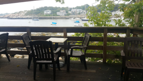 A wooden deck with chairs and a table overlooking a calm bay with boats and houses in the background.