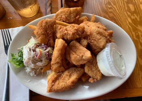 A plate of fried fish fillets with French fries, coleslaw, and a small cup of dipping sauce.