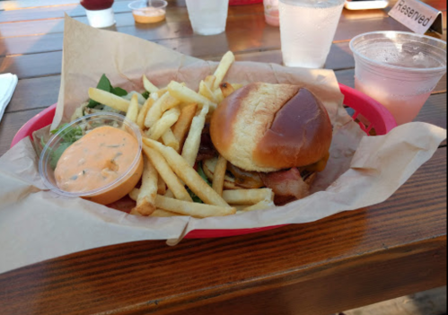 A burger with bacon on a bun, served with fries and a dipping sauce, on a wooden table.
