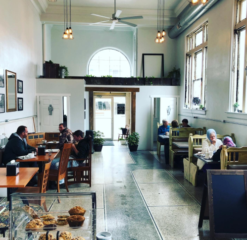 A cozy café interior with patrons seated at tables and benches, large windows, and a display case of baked goods.