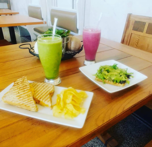 A wooden table with two smoothies, a plate of chips, and a sandwich on a white dish.