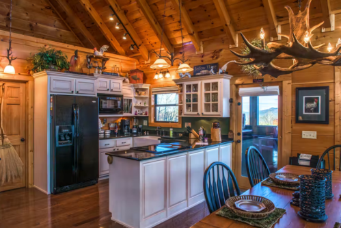 Cozy wooden kitchen with a large island, black appliances, and a rustic dining area featuring a wooden table and antler chandelier.