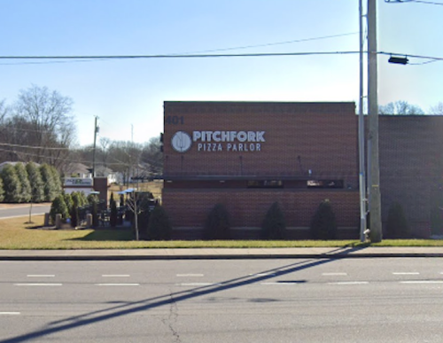 Exterior view of Pitchfork Pizza Parlor, a brick building with a sign, surrounded by grass and trees.