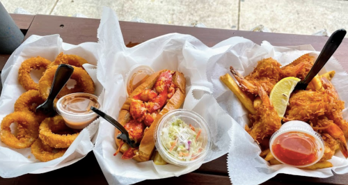 A platter featuring onion rings, a lobster roll with coleslaw, and fried shrimp with dipping sauce.