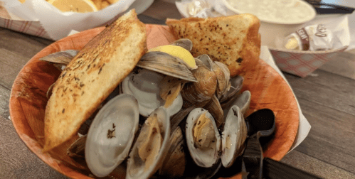 A bowl of clams with lemon and toast, served on a wooden table, with a side of soup in the background.