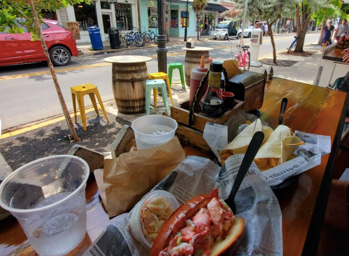 A table with a lobster roll, coleslaw, and drinks, set in a vibrant outdoor dining area with colorful chairs.