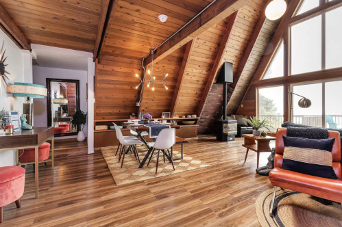 Cozy, modern living space with wooden beams, a dining area, and stylish furniture, featuring large windows and warm lighting.