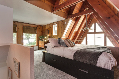 Cozy A-frame bedroom with wooden beams, large windows, and a plush bed, overlooking a serene outdoor view.