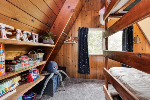 Cozy wooden cabin bedroom with bunk beds, games on a shelf, and a plush toy in a corner. Natural light from a window.